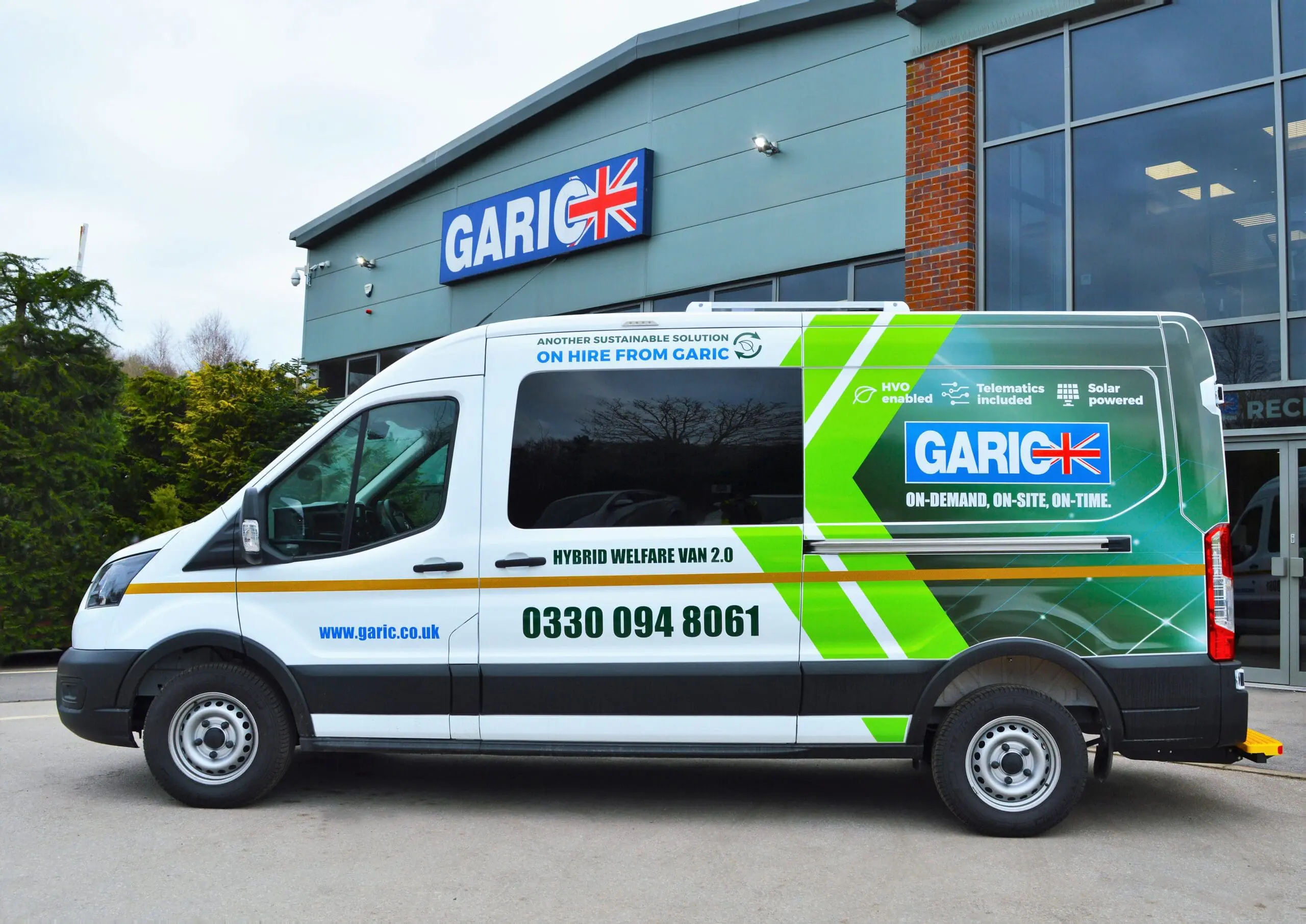 White Garic hybrid welfare van parked outside a modern building with large windows and a Garic sign.