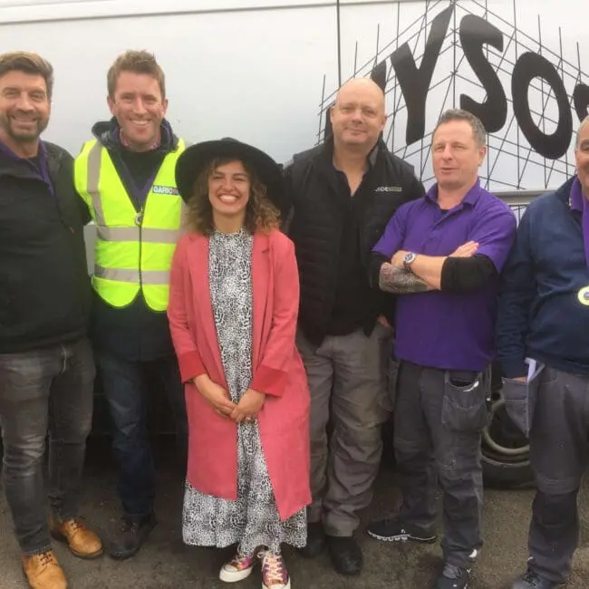 Six people stand smiling in front of a white van, one woman in a pink coat among five men.
