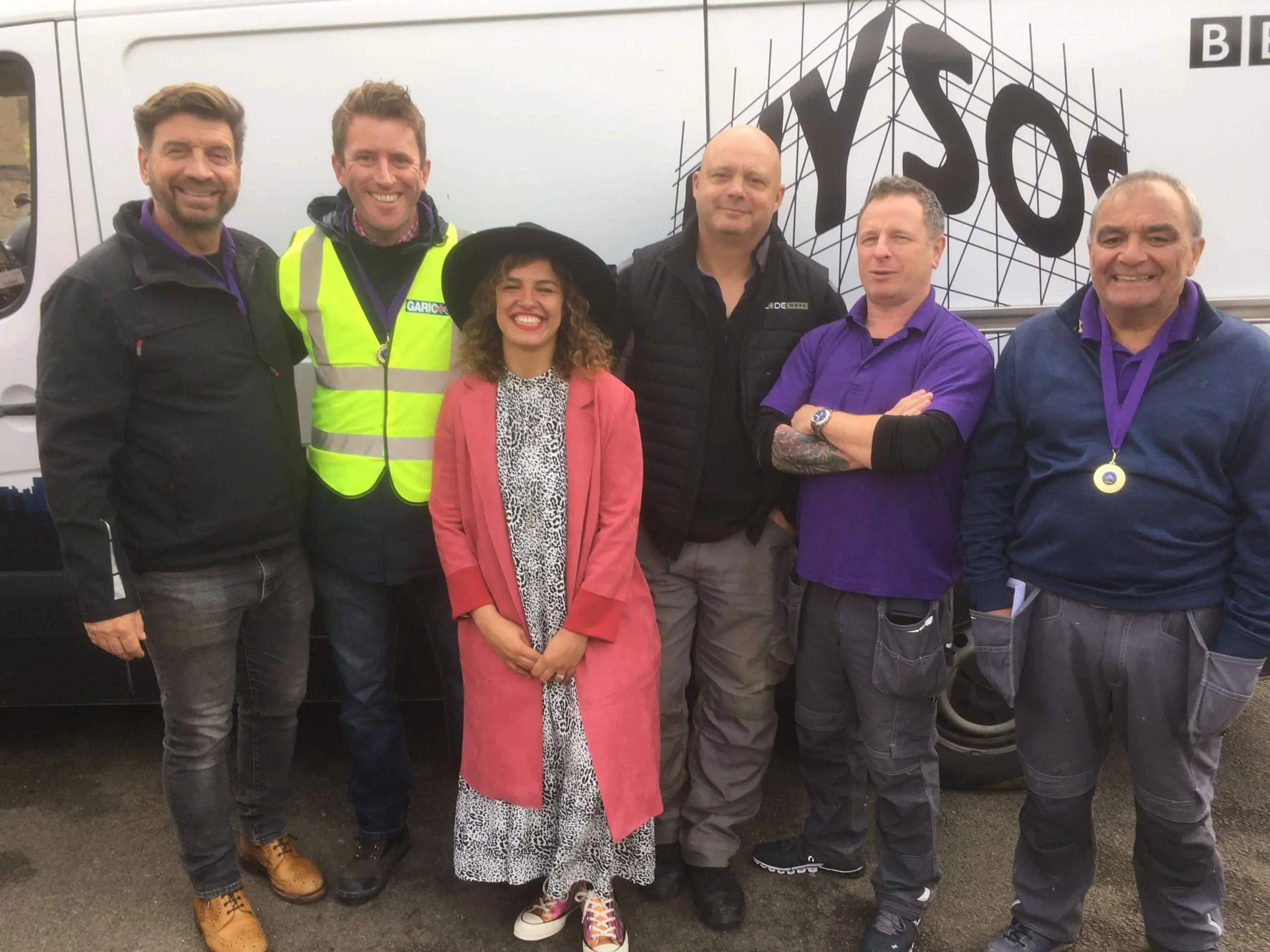 Six people stand smiling in front of a white van, one woman in a pink coat among five men.