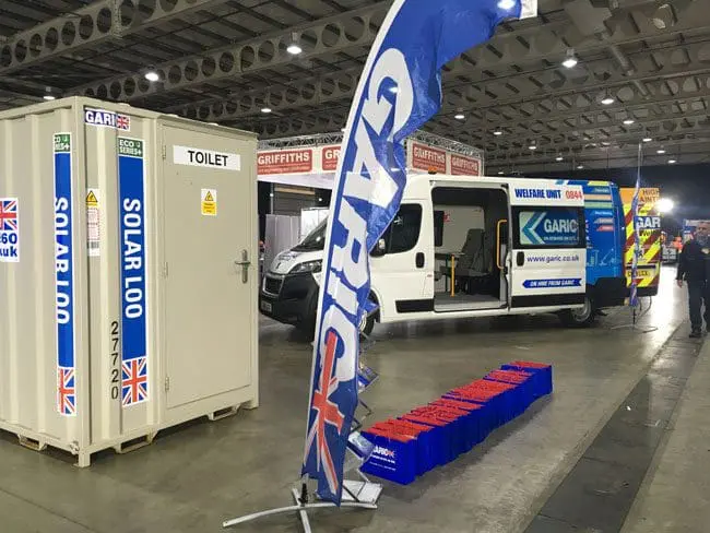 Indoor exhibition with a portable solar toilet, Garic van, promotional flags, and giveaway bags on the floor.
