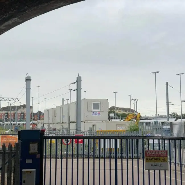 Industrial site behind a blue gate and fence, with warning signs and portable buildings in the background.