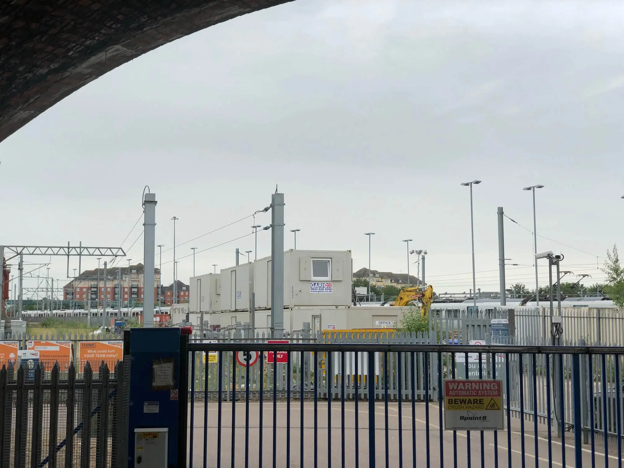 Industrial site behind a blue gate and fence, with warning signs and portable buildings in the background.