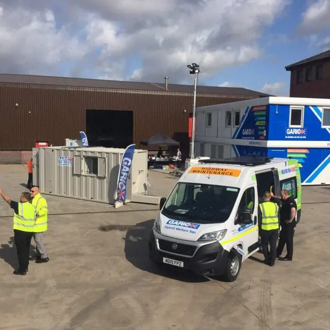 A maintenance van and workers in high-visibility vests at an industrial site with buildings and containers.