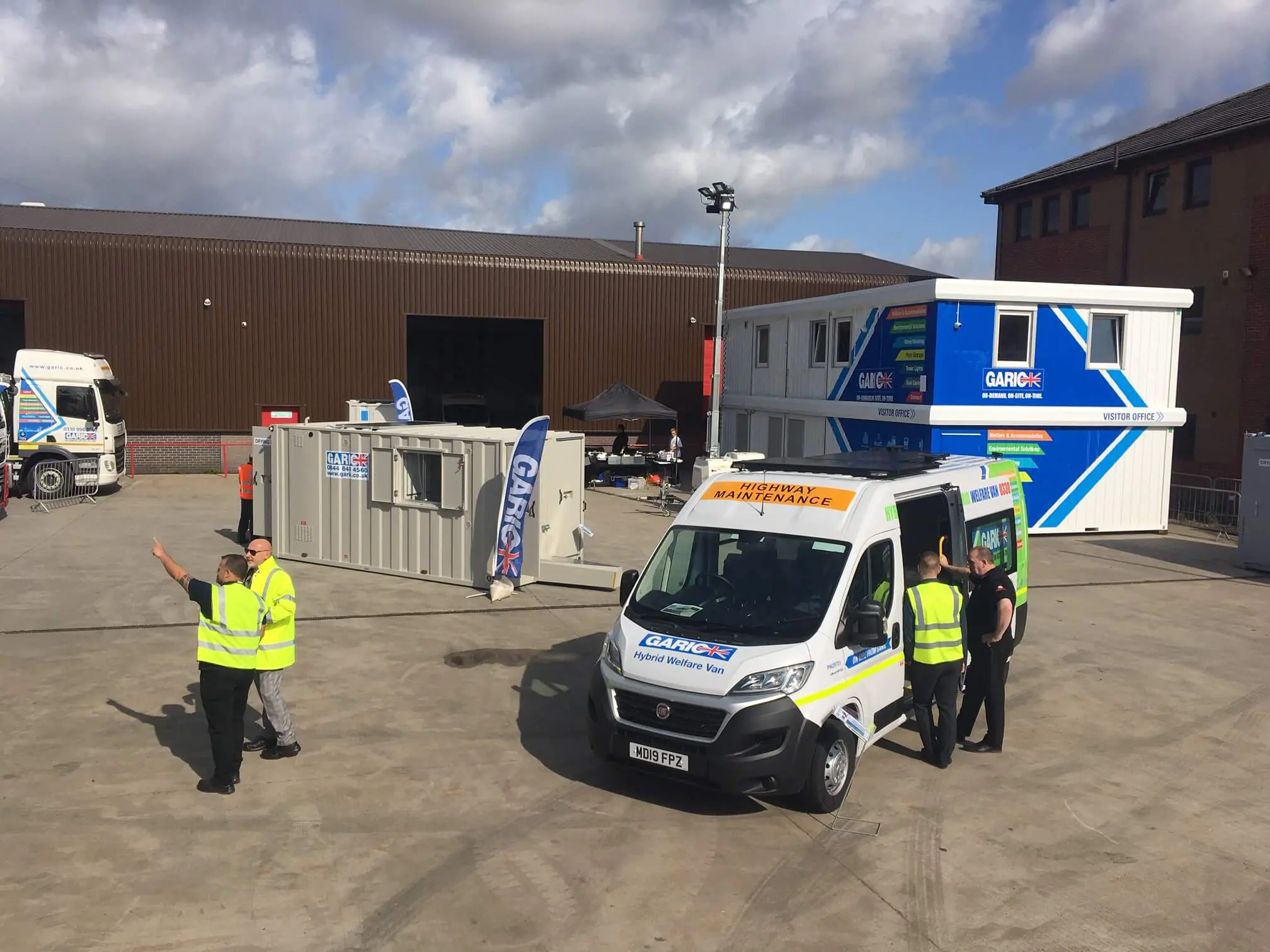 A maintenance van and workers in high-visibility vests at an industrial site with buildings and containers.