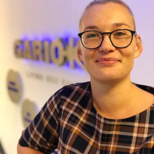 Smiling person with glasses in a plaid shirt stands indoors near a blurred sign on the wall.