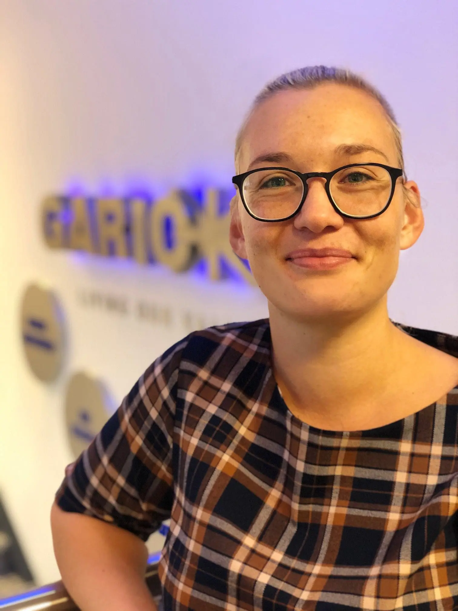 Smiling person with glasses in a plaid shirt stands indoors near a blurred sign on the wall.