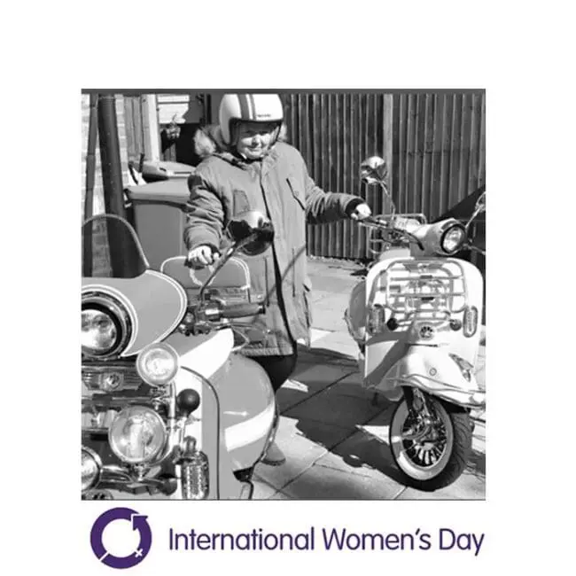 Woman in a helmet stands beside two parked scooters. International Women’s Day logo and text below.