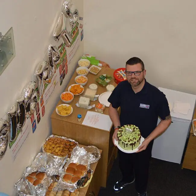 Man holding a cake stands by a table with food and “congrats” balloons on the wall.