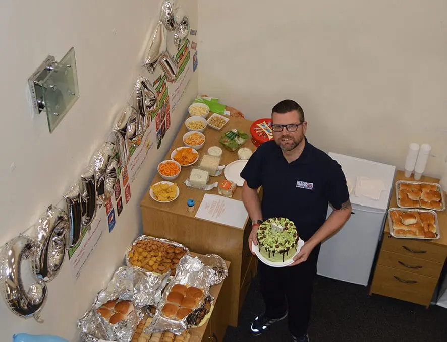 Man holding a cake stands by a table with food and “congrats” balloons on the wall.