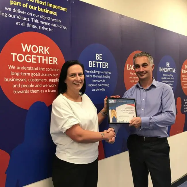 Two people shaking hands as one receives a certificate, standing in front of a wall with business values.