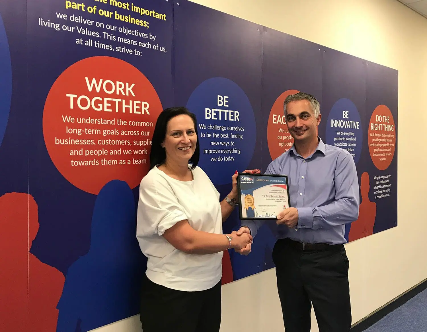 Two people shaking hands as one receives a certificate, standing in front of a wall with business values.