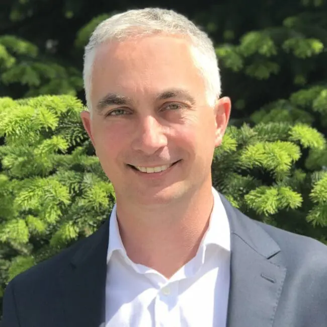 Smiling man in a suit jacket stands outdoors in front of green pine tree branches.