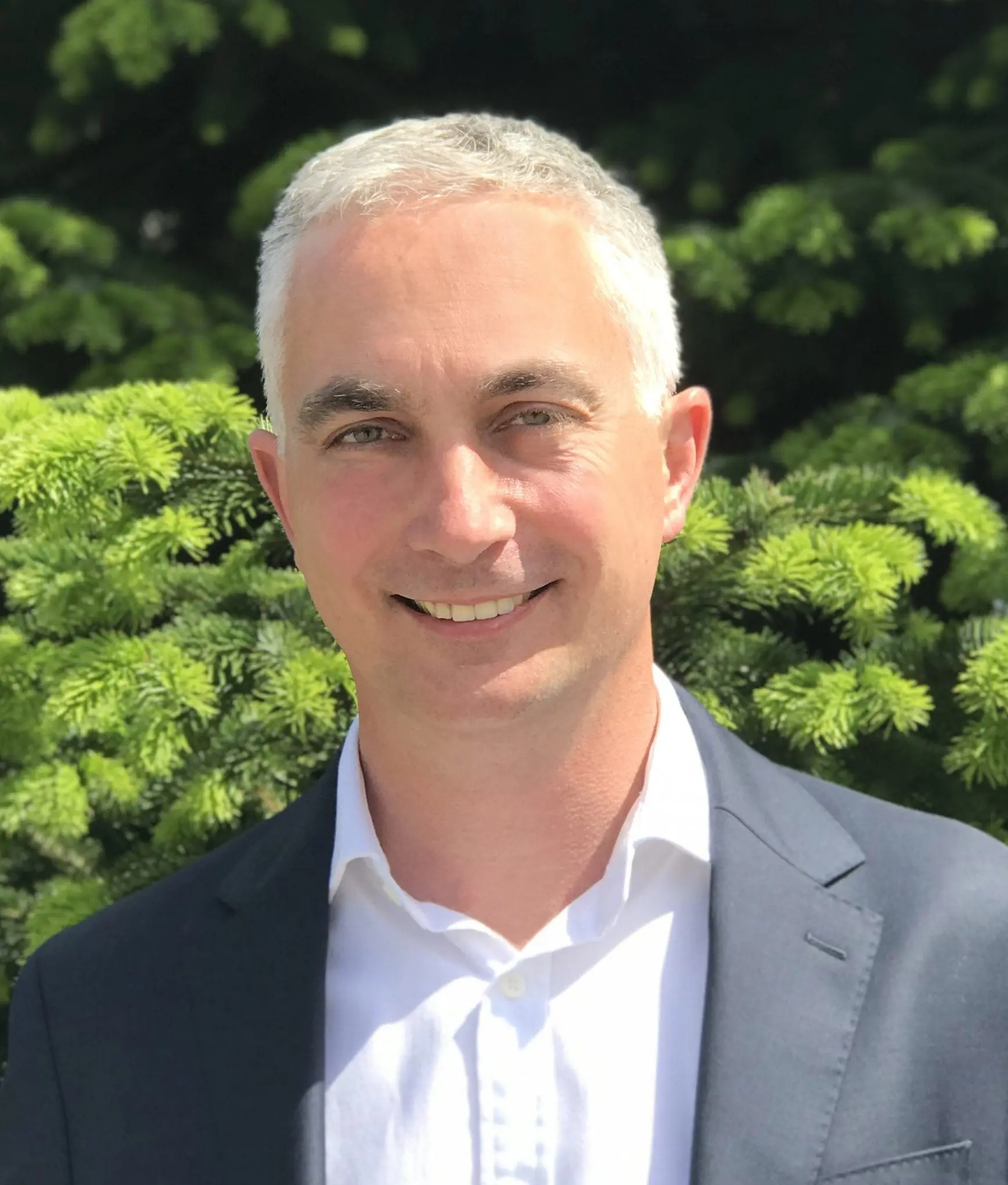 Smiling man in a suit jacket stands outdoors in front of green pine tree branches.