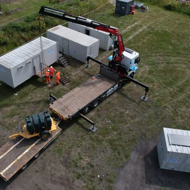 A crane truck lifts equipment from a trailer near white containers and workers in orange safety gear.