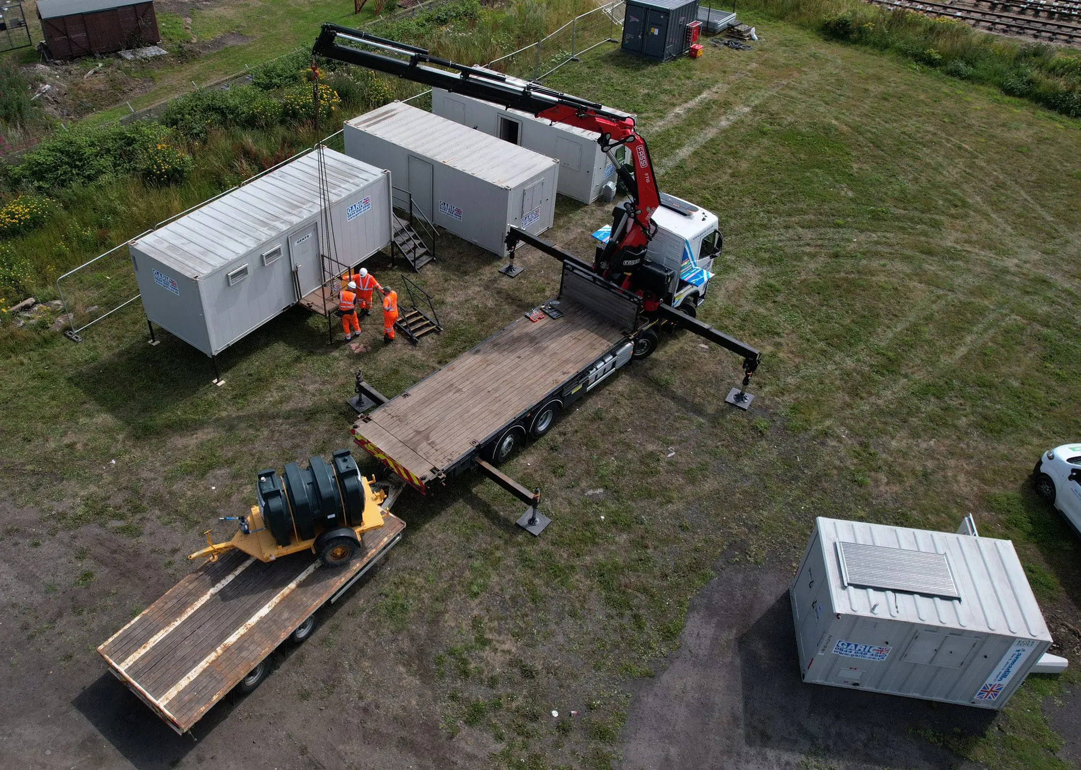 A crane truck lifts equipment from a trailer near white containers and workers in orange safety gear.
