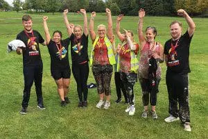 Seven people stand on grass with arms raised, smiling, wearing medals and sportswear, celebrating outdoors.