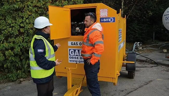 Two workers in safety gear talk beside a yellow gas oil machine outdoors, one with the equipment panel open.