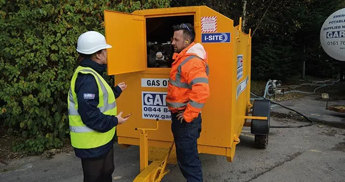 Two workers in safety gear talk beside a yellow gas oil machine outdoors, one with the equipment panel open.