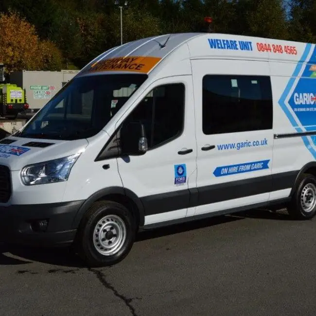 White welfare van with blue and yellow graphics parked outdoors in a lot with trees and equipment in the background.