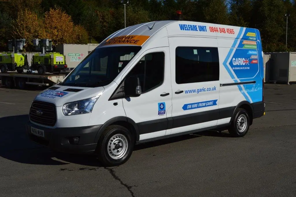 White welfare van with blue and yellow graphics parked outdoors in a lot with trees and equipment in the background.