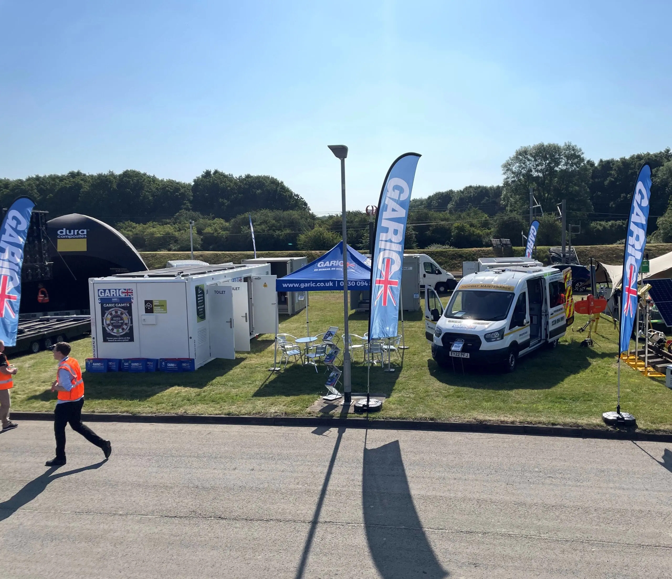 Outdoor event setup with GARIC banners, tents, vehicles, and people on a sunny day.