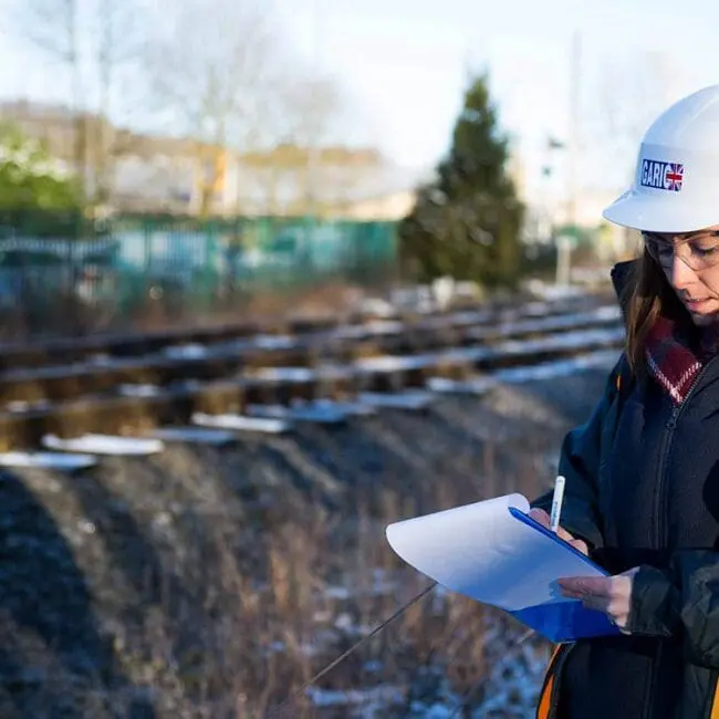 Woman in safety gear writes on a clipboard by railway tracks on a sunny, winter day.