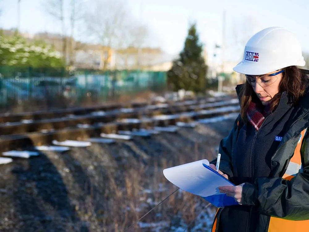 Woman in safety gear writes on a clipboard by railway tracks on a sunny, winter day.