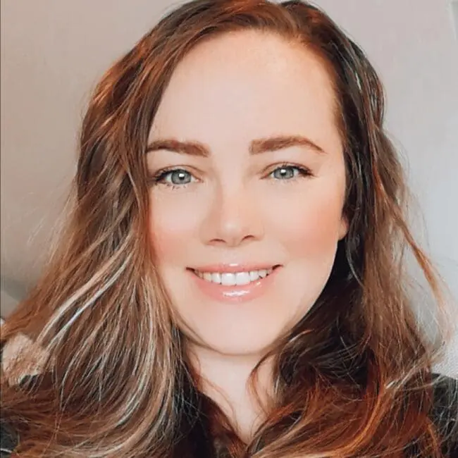 A woman with long wavy brown hair smiles at the camera against a neutral background.