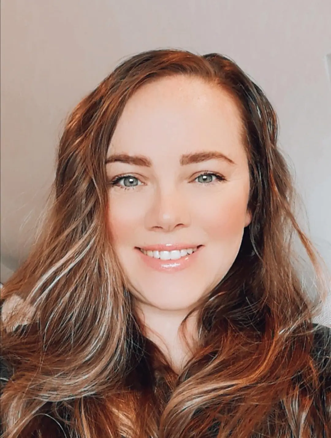 A woman with long wavy brown hair smiles at the camera against a neutral background.