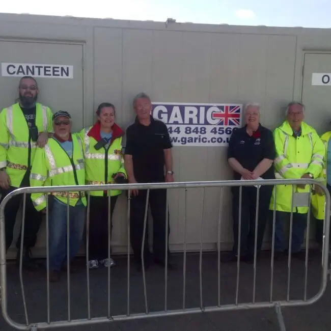 Seven people in hi-vis clothing stand smiling outside portable toilet, canteen, and office units behind a barrier.