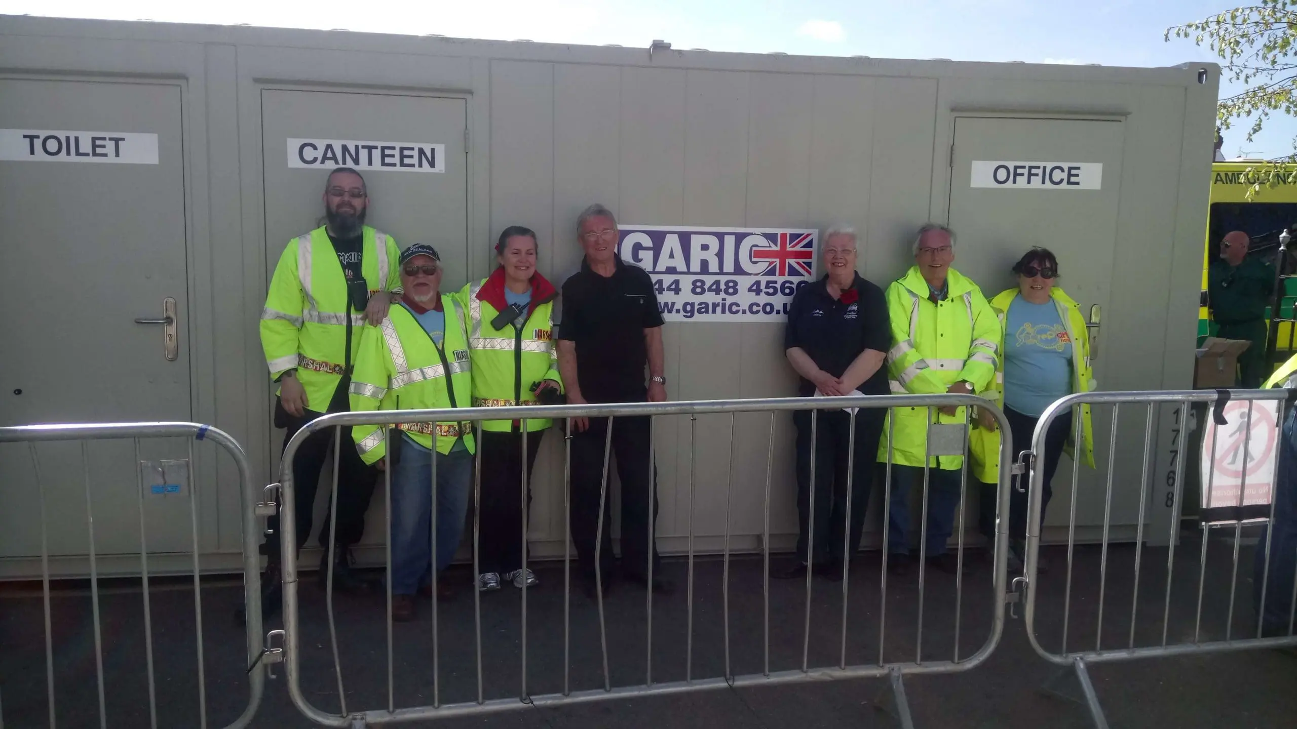 Seven people in hi-vis clothing stand smiling outside portable toilet, canteen, and office units behind a barrier.