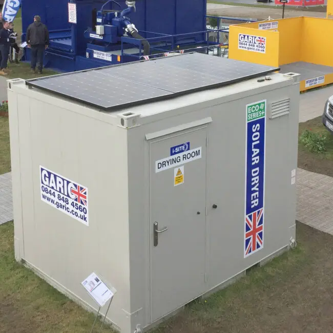 Small white drying room with solar panels on top, labeled Solar Dryer, at an outdoor exhibition.
