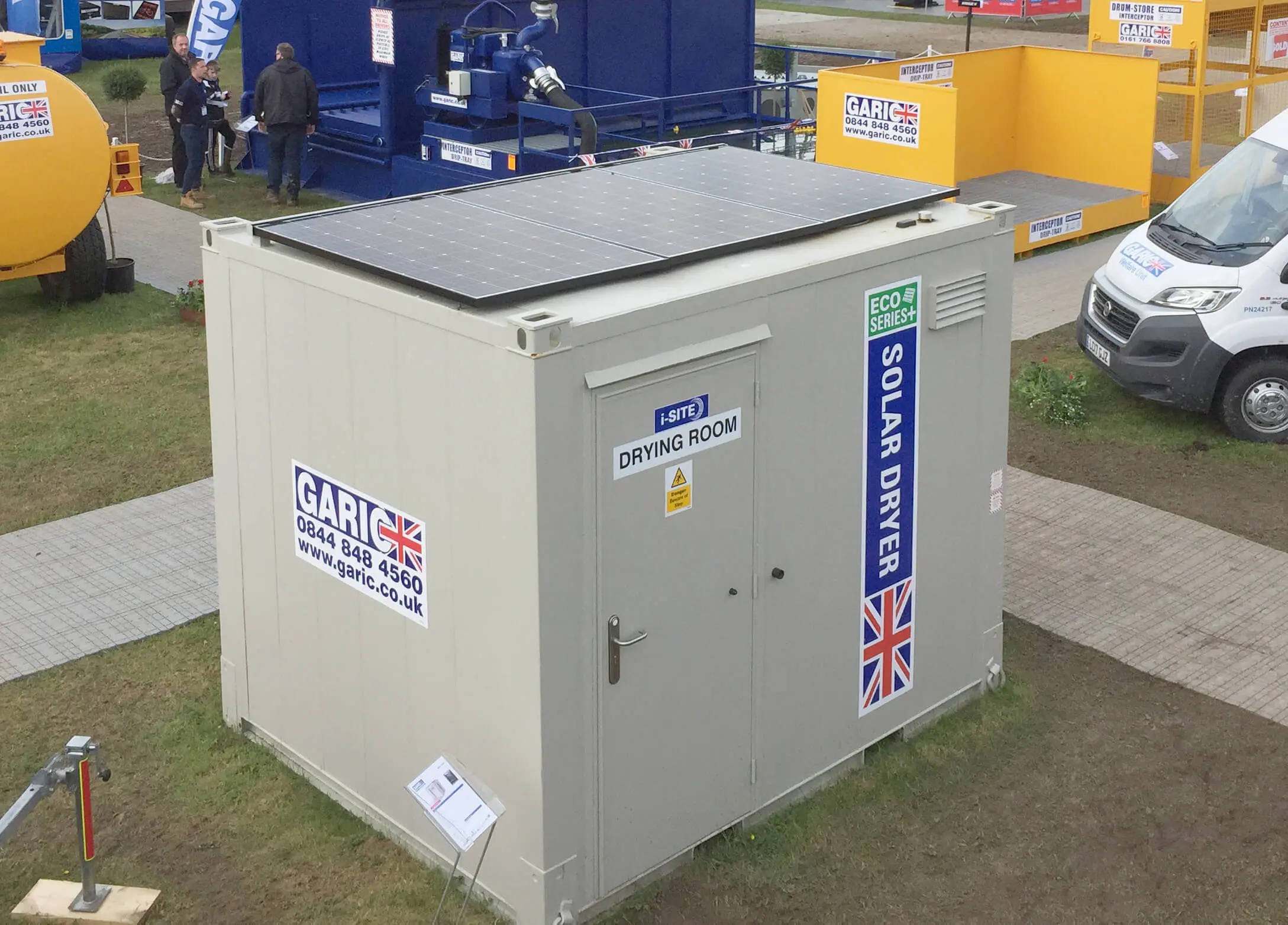 Small white drying room with solar panels on top, labeled Solar Dryer, at an outdoor exhibition.