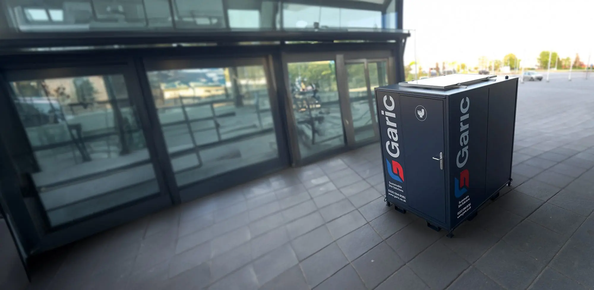 A Solar Loo 8 sanitation station stands on tiled pavement near a glass building entrance under the sun.