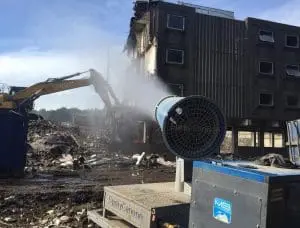 A machine sprays water as an excavator demolishes a multi-story building on a construction site.