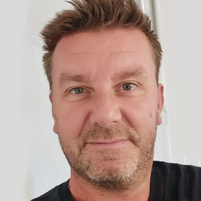 Middle-aged man with short brown hair and light stubble, wearing a dark shirt, looking at the camera.