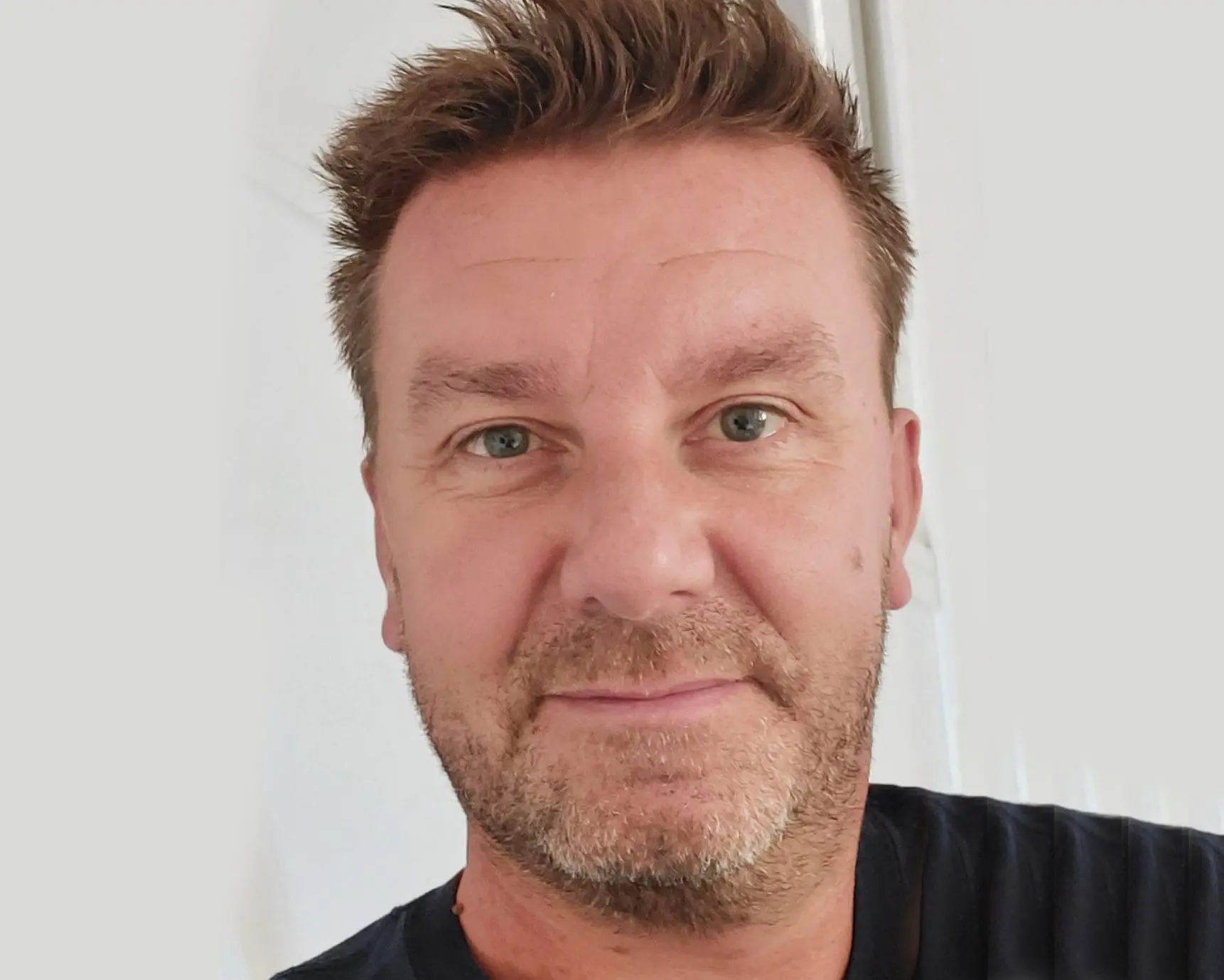 Middle-aged man with short brown hair and light stubble, wearing a dark shirt, looking at the camera.