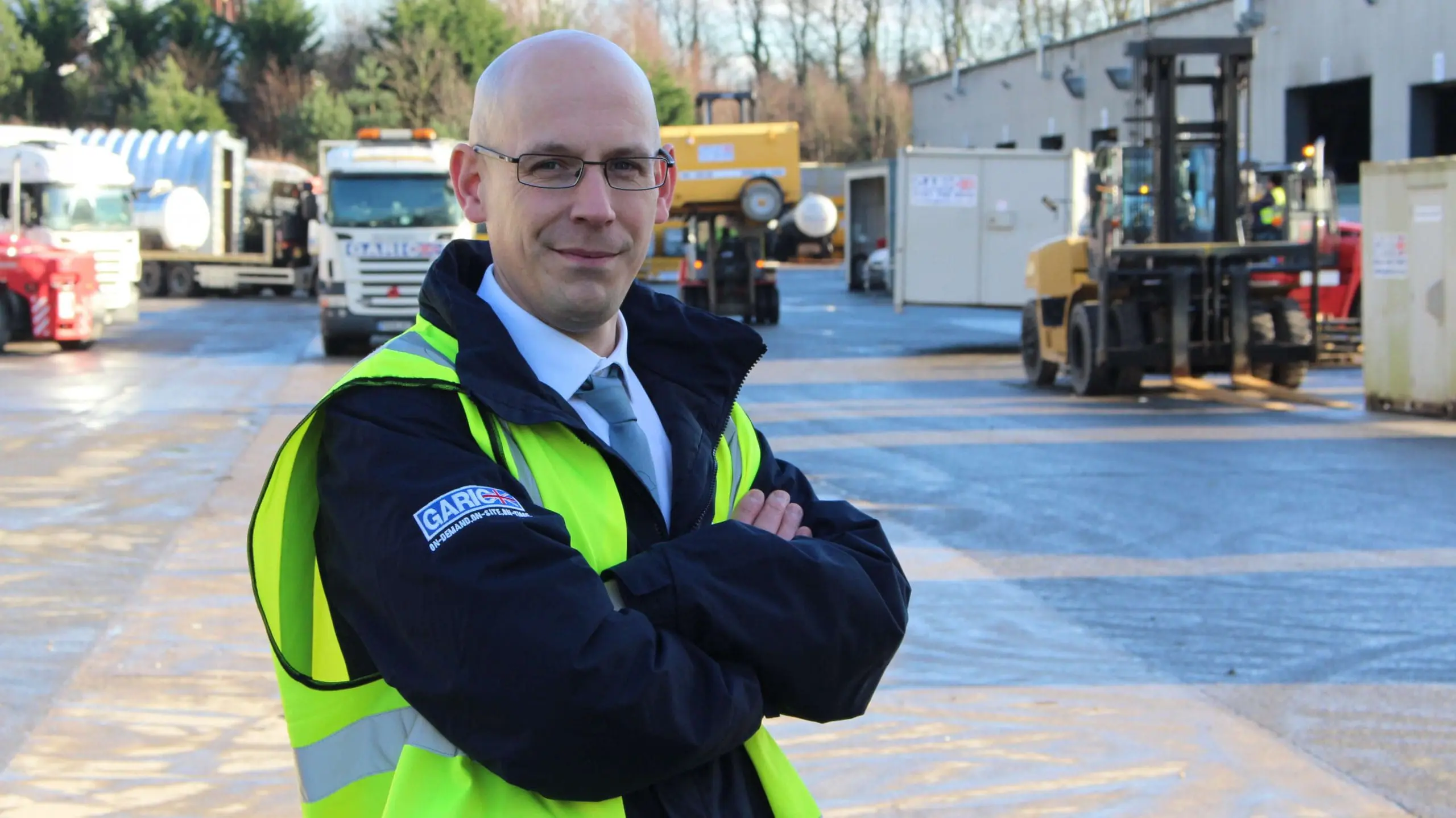 Bald man in a yellow safety vest stands with arms crossed at an industrial worksite with trucks and equipment.