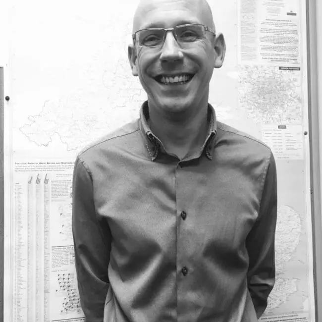 A smiling man in glasses stands in front of a map display on a wall. Black and white photo.