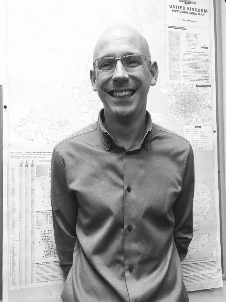A smiling man in glasses stands in front of a map display on a wall. Black and white photo.