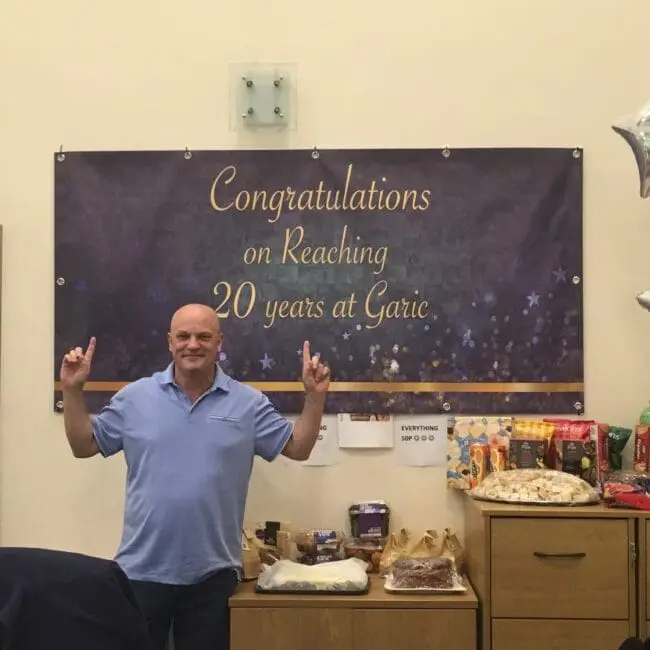 A man smiles and points up at a banner that says Congratulations on Reaching 20 years at Garic.