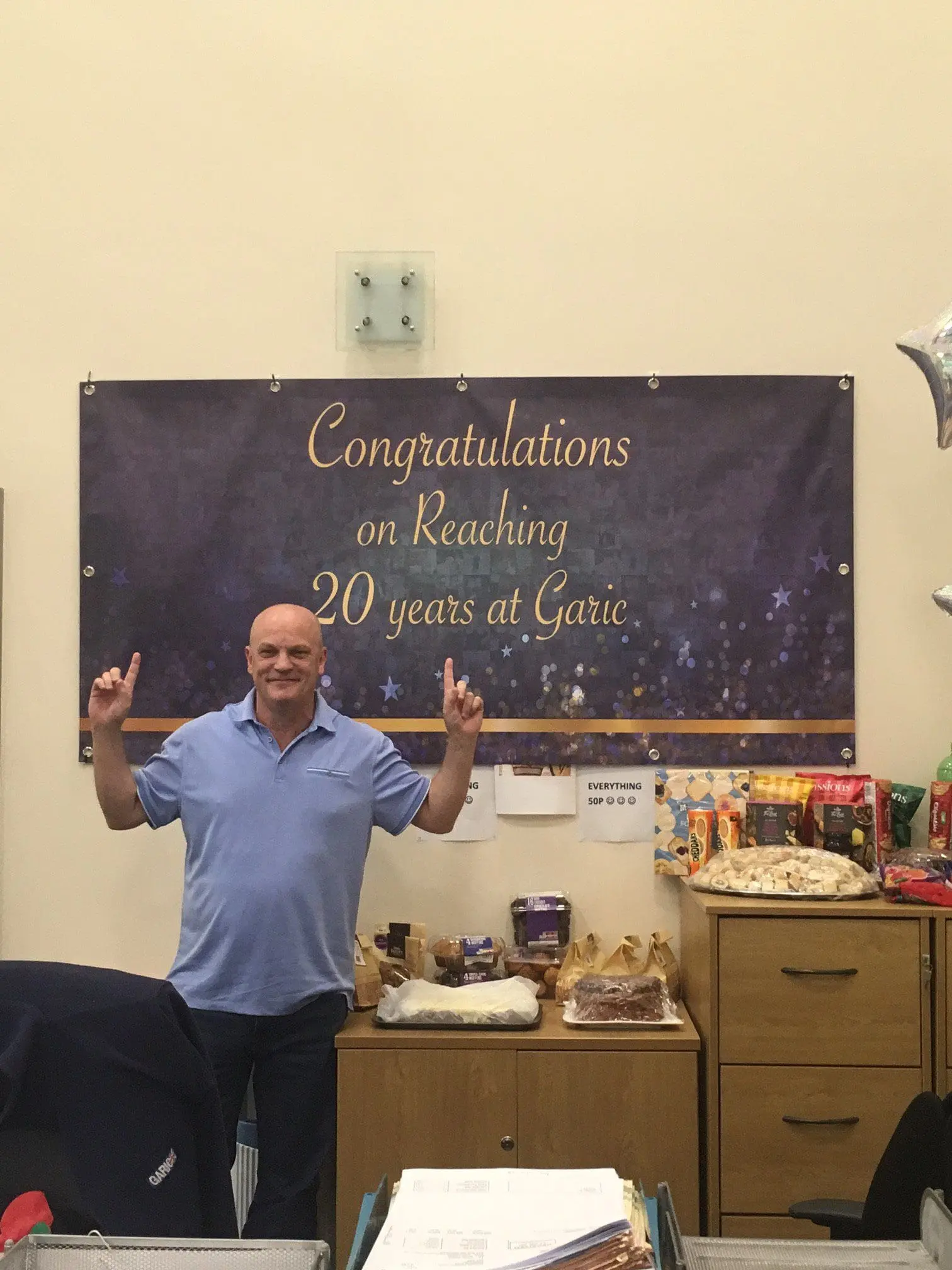 A man smiles and points up at a banner that says Congratulations on Reaching 20 years at Garic.