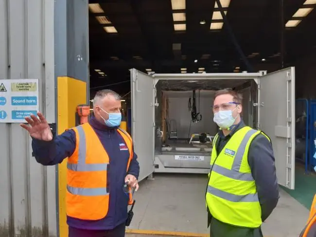 Two men in safety vests and masks talk outside an industrial building with open doors behind them.