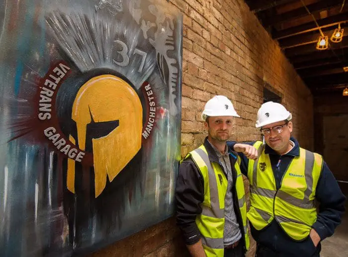 Two men in hard hats and vests stand beside a Veterans Garage Manchester mural on a brick wall.
