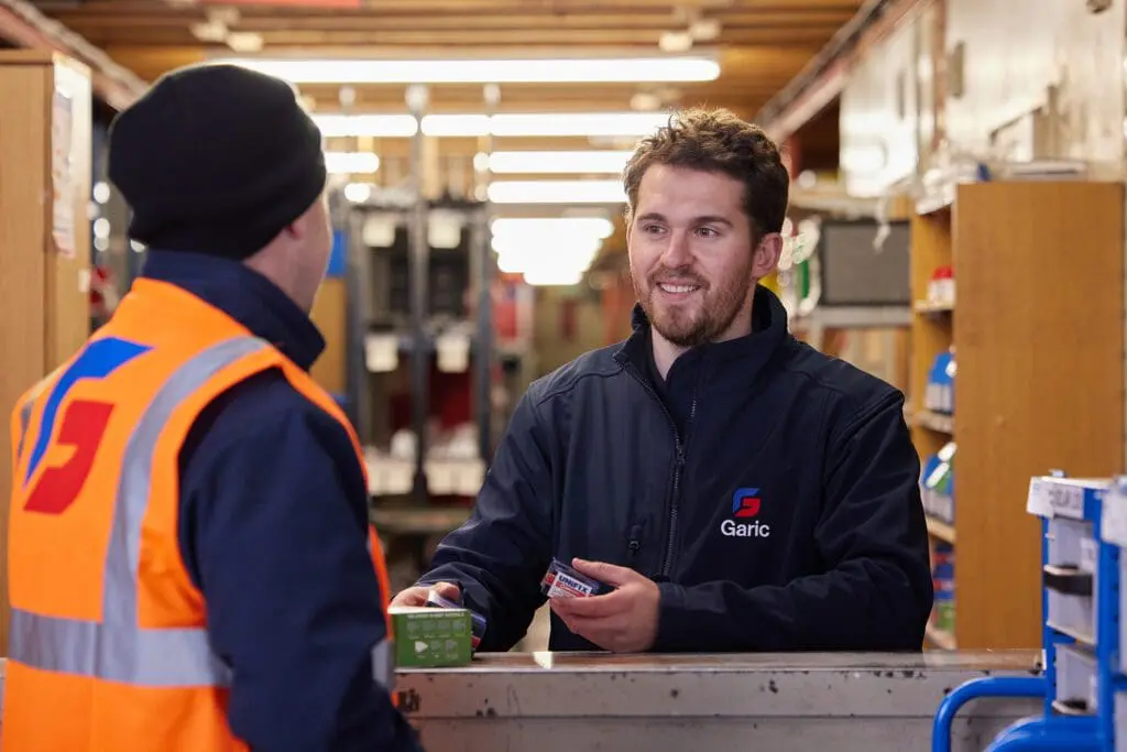 A smiling shop worker hands items to a customer in a hi-vis vest at a counter in a hardware store.