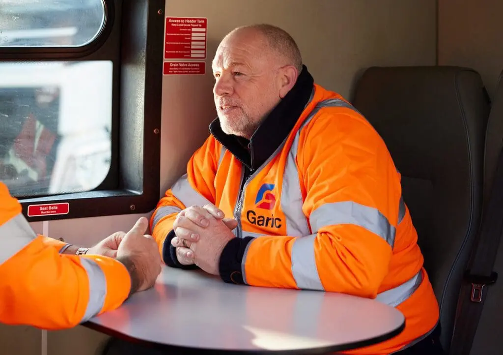 Man in an orange high-visibility jacket sits at a table, talking with another person inside a vehicle or cabin.