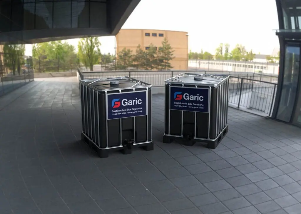 Two Waste IBC 1000 tanks are positioned on a paved outdoor area near modern buildings and trees.