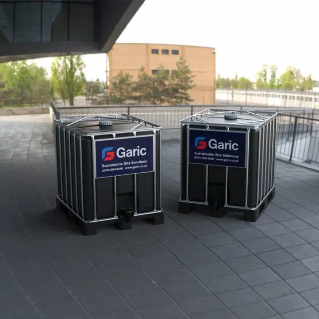 Two Waste IBC 1000 tanks are positioned on a paved outdoor area near modern buildings and trees.