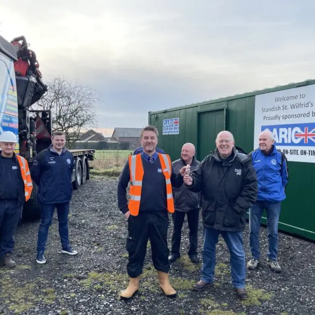 Six men in workwear stand smiling beside a truck and green container on a cloudy day.