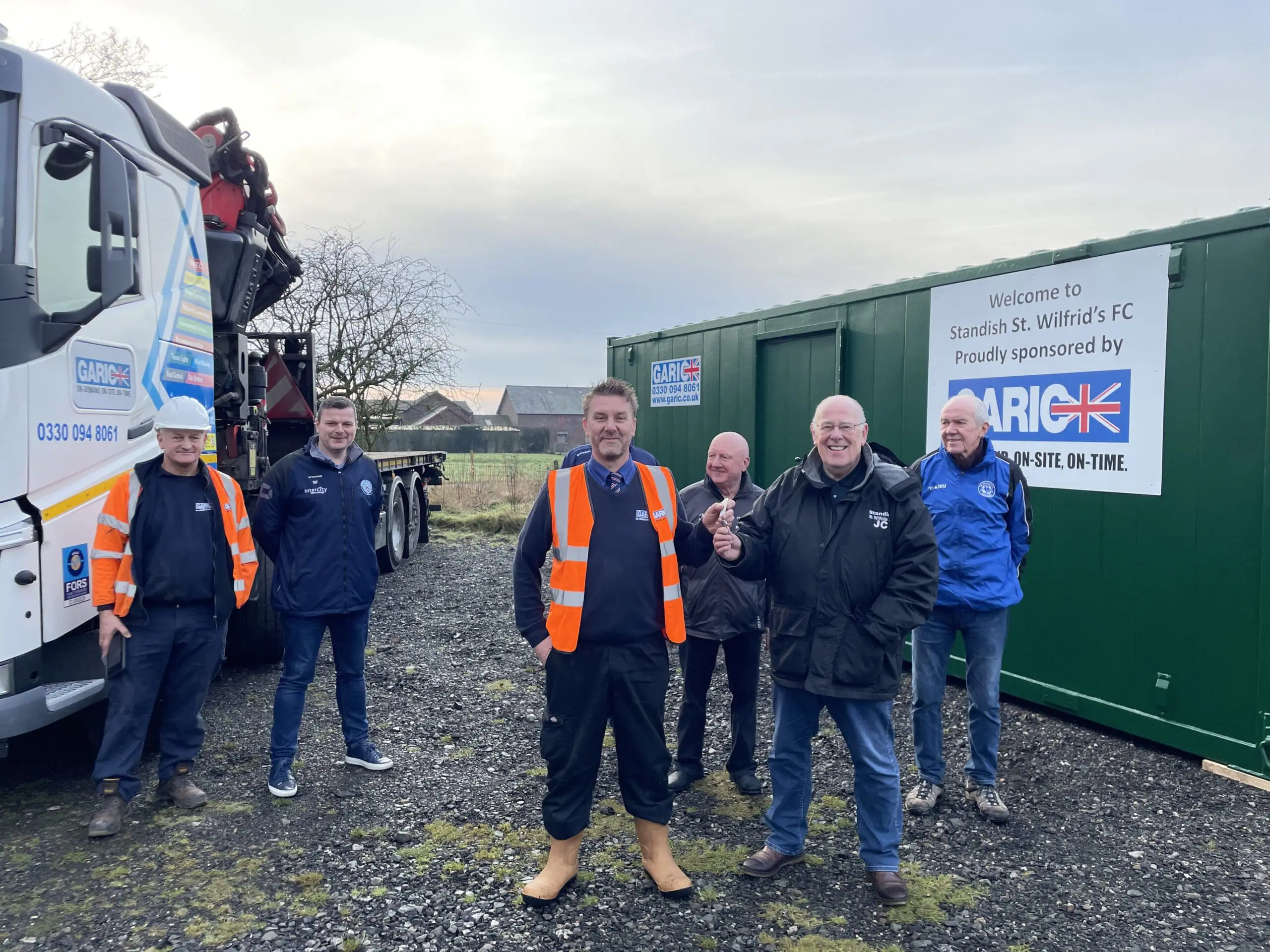 Six men in workwear stand smiling beside a truck and green container on a cloudy day.